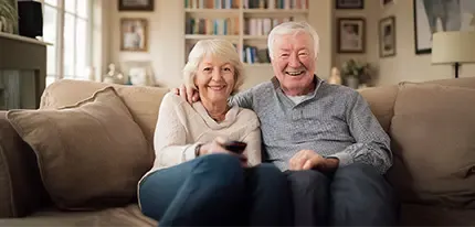 An older couple sits smiling on a couch, the woman holding a remote control.